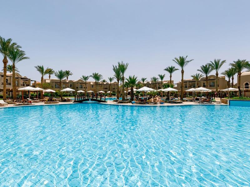 Clear blue pool surrounded by palm trees and sun umbrellas at a resort under a bright sky.