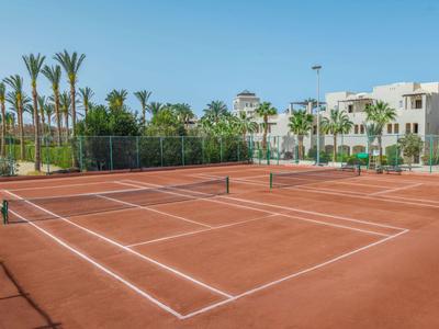 Court de tennis en terre battue entouré de palmiers et de bâtiments sous un ciel clair.