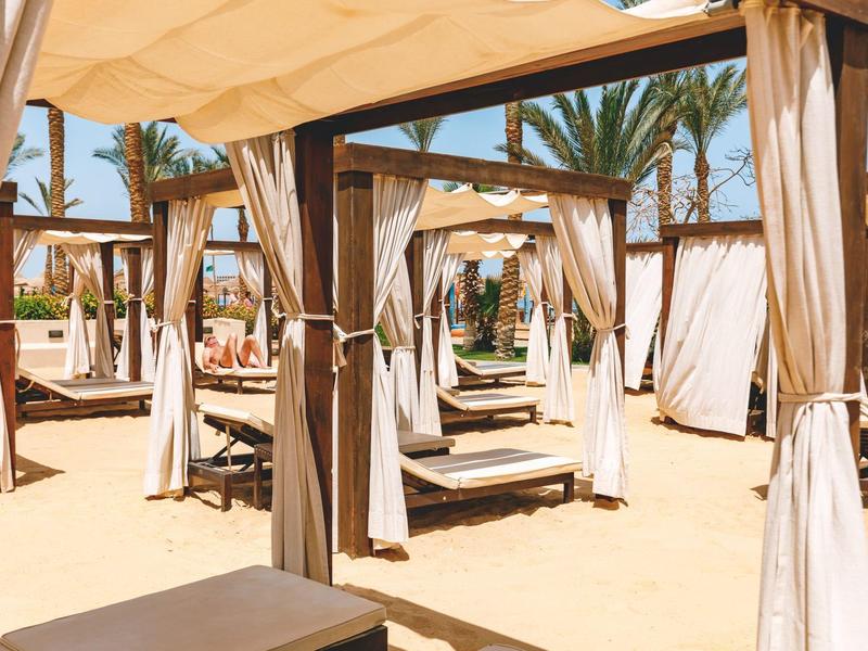 Strandcabana's met gordijnen onder schaduwdaken en palmbomen op zonnig zandstrand.