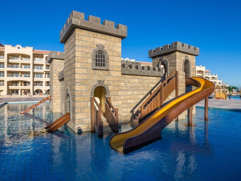 Castillo de juegos acuáticos con toboganes en una piscina junto a un hotel bajo un cielo azul claro.