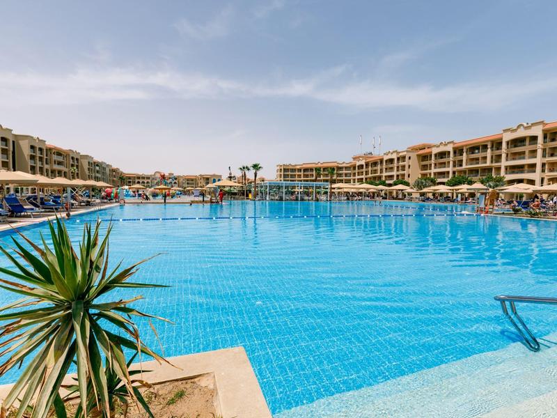 Grande piscine bleue entourée de bâtiments d'hôtel sous un ciel clair.