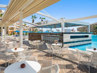 Poolside bar area with white tables and wicker chairs under shaded pergola near a blue swimming pool.