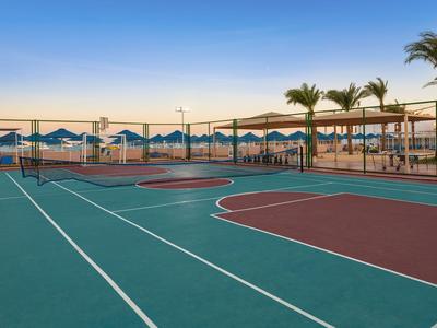 Campo sportivo vuoto con campi da tennis e basket accanto a una spiaggia con ombrelloni.