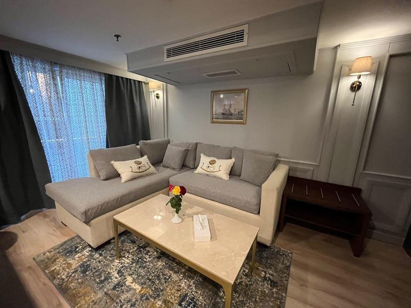 Modern hotel room with beige sectional sofa, coffee table, rug, and framed artwork.