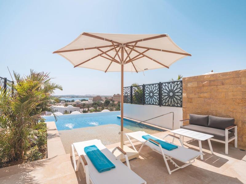 Terrazza con lettini, ombrellone e piscina con vista su edifici bianchi.