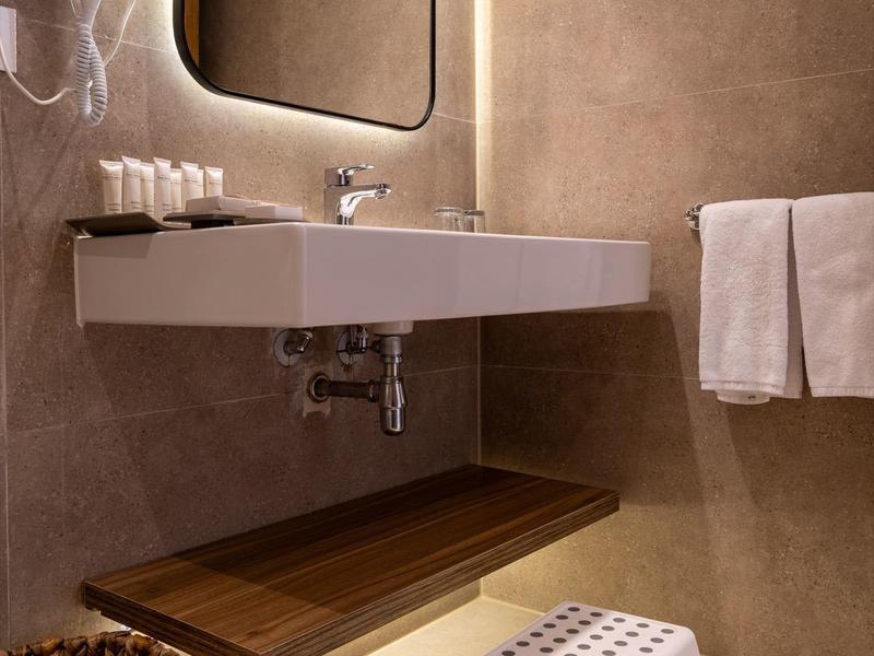 Baño moderno con espejo iluminado, lavabo de pared, toallas y taburete de plástico.