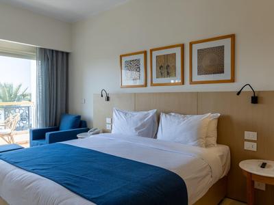 Chambre d'hôtel moderne avec lit double, couverture bleue et balcon avec vue sur les palmiers.