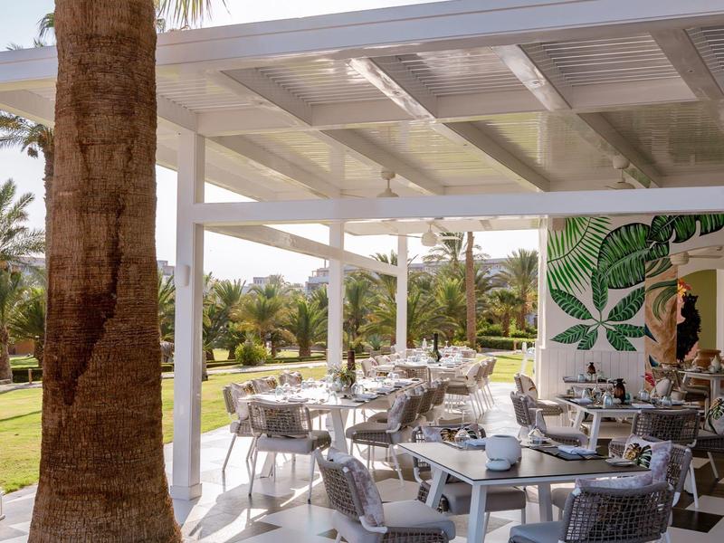 Espace extérieur d'un hôtel avec terrasse couverte, chaises et tables à côté des palmiers.