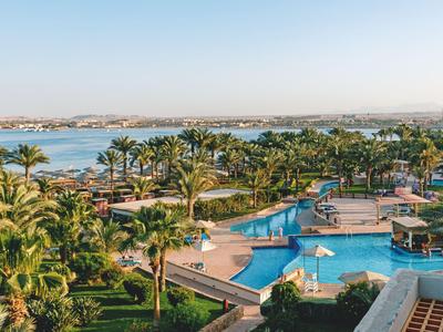 Großer Pool mit Sonnenschirmen, umgeben von Palmen und Blick auf das Meer.