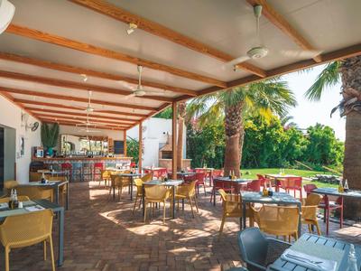 Overdekt restaurantterras met tafels en stoelen, omgeven door palmbomen en tuin.