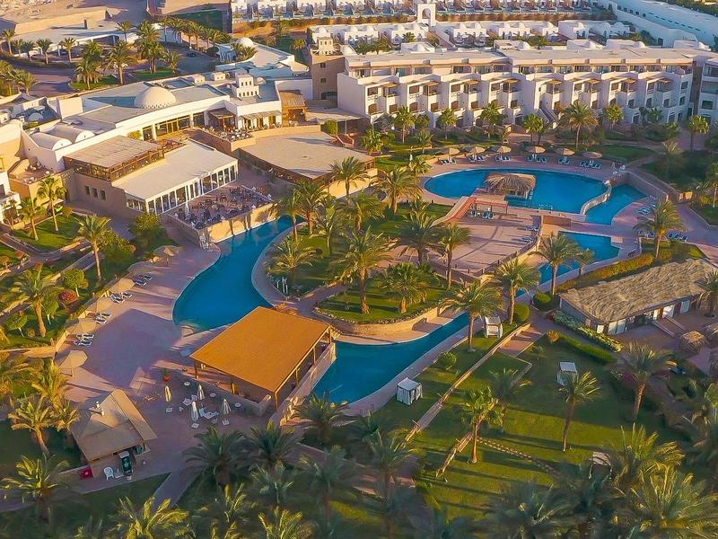 Vista aérea de un resort con piscinas, palmeras y amplios jardines al atardecer.