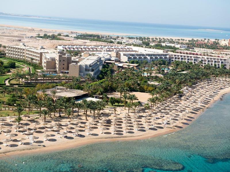 Aerial view of a large beachfront resort with umbrellas and sunbeds along the sandy shore near clear blue water.