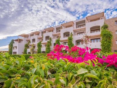 Modern hotel with balconies, pink flowers, green plants, and a partly cloudy sky.