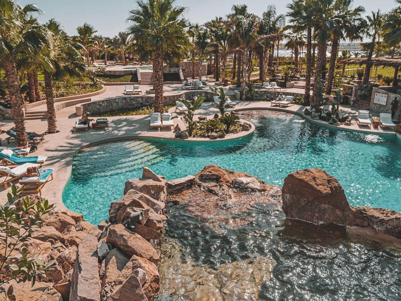 Large pool area with rocks, palm trees, and lounge chairs in a sunny hotel resort.