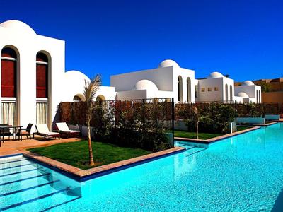 White Mediterranean hotel buildings with a clear blue pool under a bright blue sky.