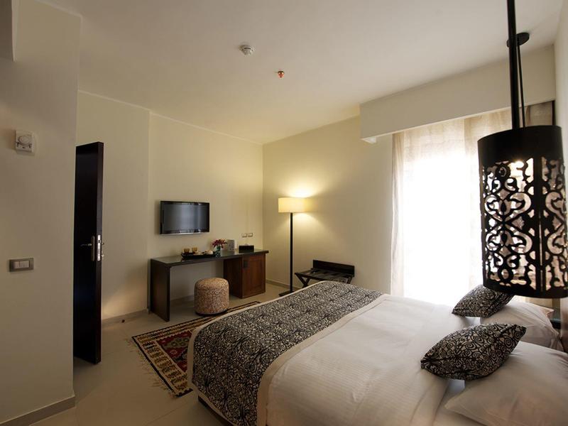 Cozy hotel room with double bed, patterned cushions, TV, lamp, and balcony door letting in natural light.