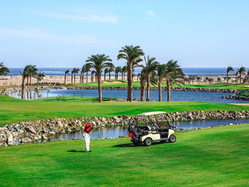 Campo de golf junto al mar con palmeras, jugador practicando y carrito de golf en césped verde.