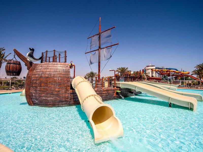 Water park with slides and pirate ship in clear blue pool under bright blue sky.