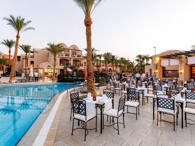 Venkovní restaurace s bazénem a řadami stolů v tropickém hotelovém resortu při západu slunce.