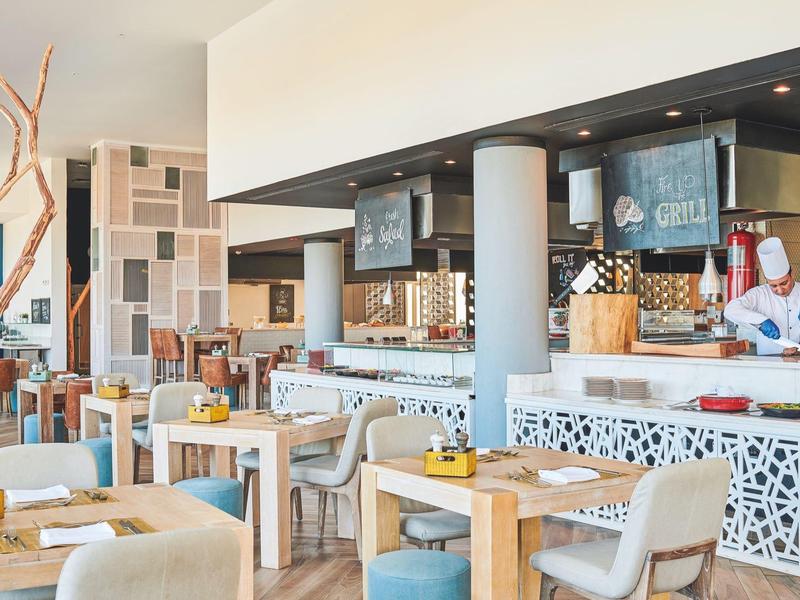 Modernes Restaurant mit Holz- und gepolsterten Stühlen, hellen Tischen und offener Bar.