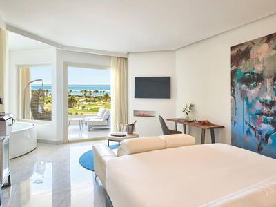 Bright hotel room with large bed, seating area, balcony, and sea view.