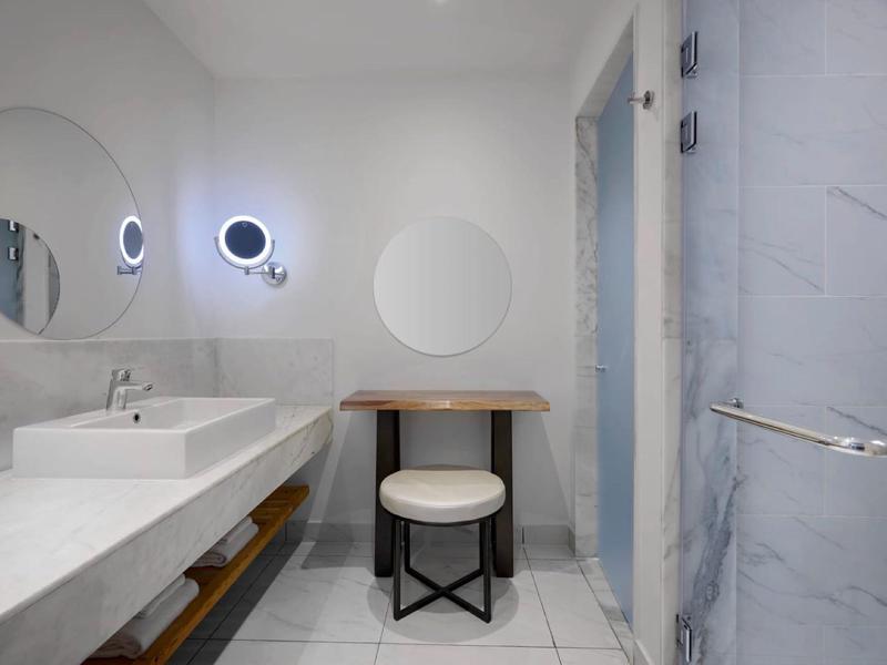 Moderne badkamer met witte marmerlook, dubbele wastafels, verlicht spiegel en kruk voor een kleine tafel.