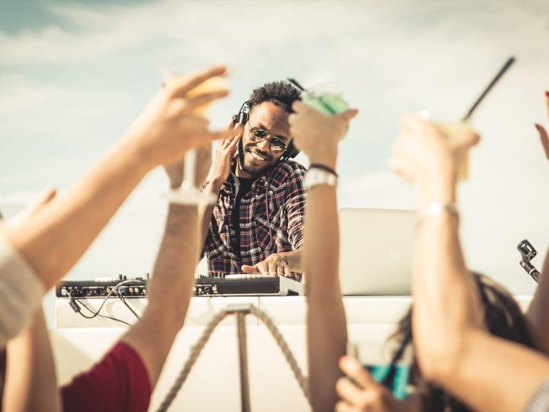 DJ plays music at an outdoor party, people dance with raised hands.
