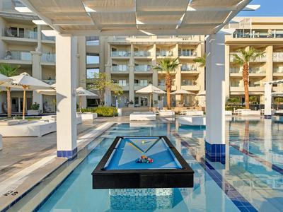 Área de piscina moderna con una mesa de billar suspendida sobre el agua en un hotel.