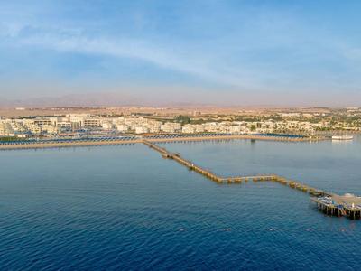 Aerial view of a long pier leading to a small pavilion over the sea, with a city and mountains in the background.