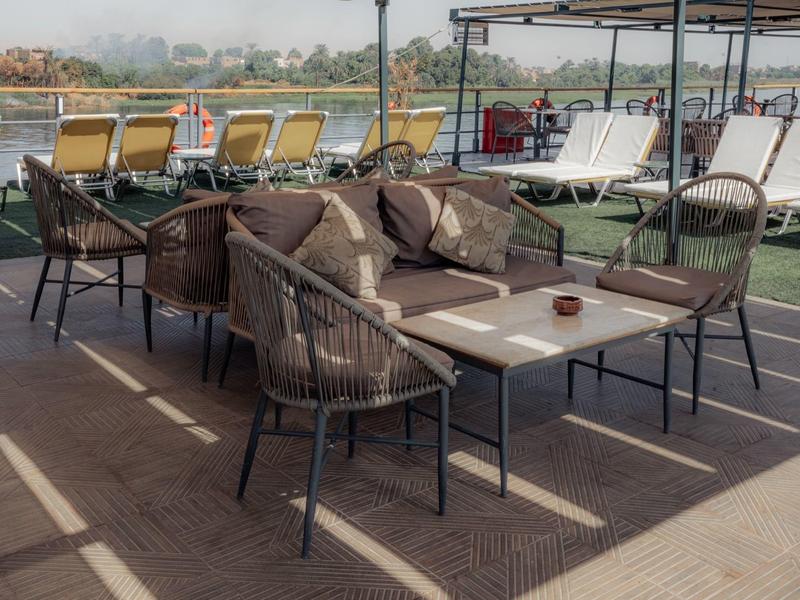 Outdoor-Lounge mit braunen Stühlen, Sofa, Tisch und Liegestühlen am Flussufer bei sonnigem Wetter.