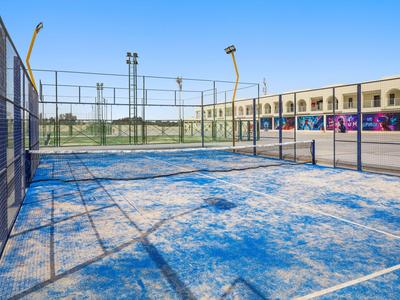 Leerer blauer Paddle-Tennisplatz mit Gitterzäunen und Gebäuden im Hintergrund.