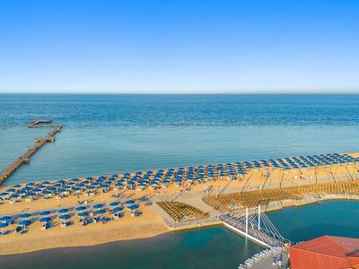 Spiaggia con ombrelloni blu e lettini accanto a un lungo molo e mare calmo.