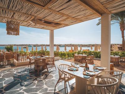 Area pranzo aperta con tavoli e sedie in legno, vista sulla piscina e sul mare sullo sfondo.