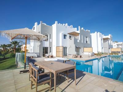 Moderner weißer Wohnkomplex mit Swimmingpool, Terrasse, Tischen, Stühlen und Sonnenschirm bei klarem Himmel.