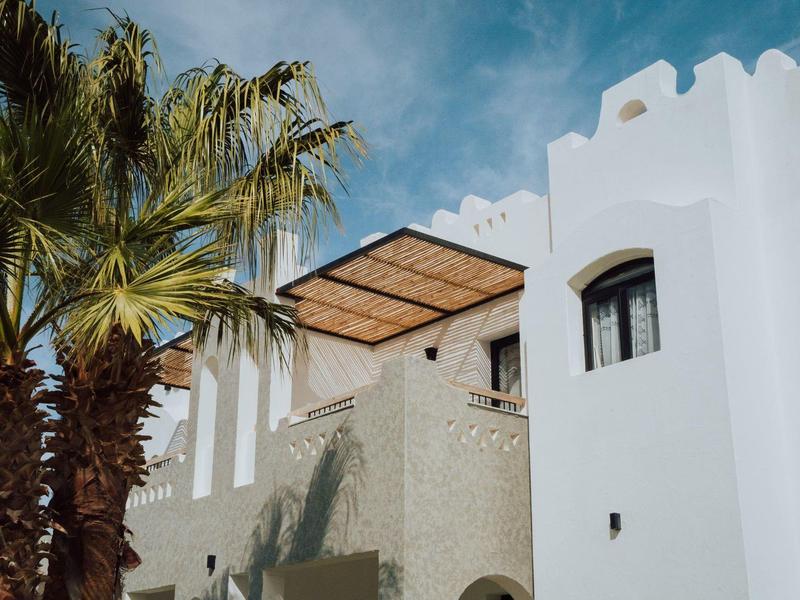 White hotel building with palm trees and a shaded balcony under blue sky.