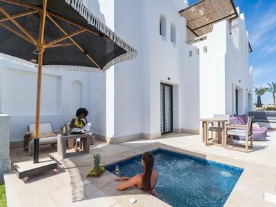 Bungalow de vacaciones blanco moderno con piscina pequeña, sombrilla y dos huéspedes relajados en el área exterior soleada.