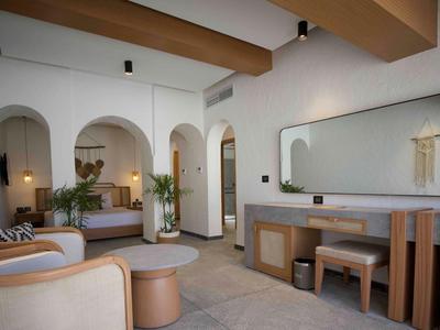 Habitación de hotel moderna con muebles de madera clara, paredes beige y sillones cómodos bajo vigas de madera.