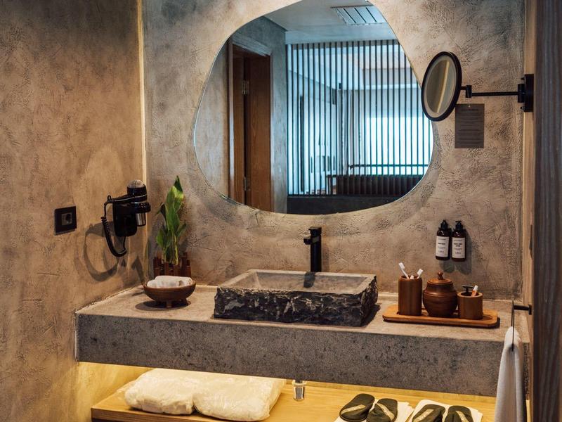Modern hotel bathroom with stone sink, circular mirror, and wooden shelf with towels and slippers.