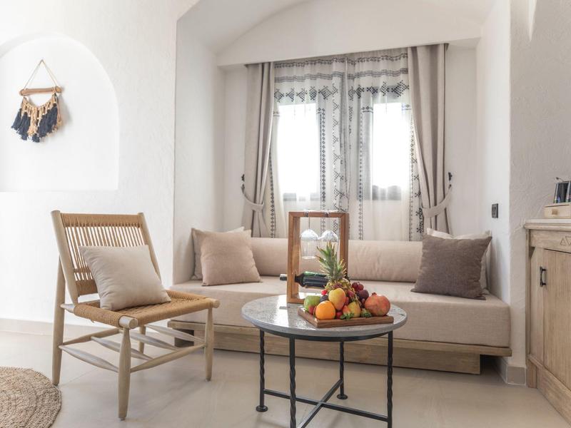 Bright hotel lounge with wooden chair, beige sofa, and round table holding fruit bowl.