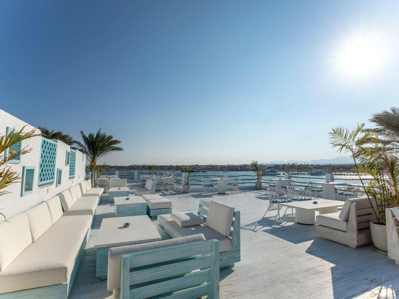 Terrazza con divani e tavoli vicino a una marina sotto un cielo soleggiato.