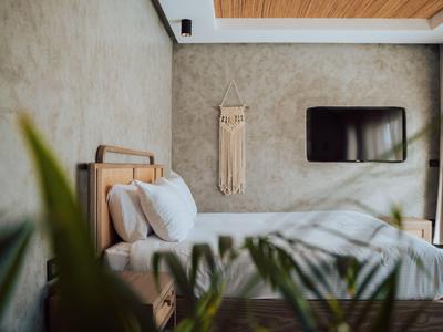 Modern hotel room with wooden bed, white linens, decorative wall hanging, and TV on concrete wall.
