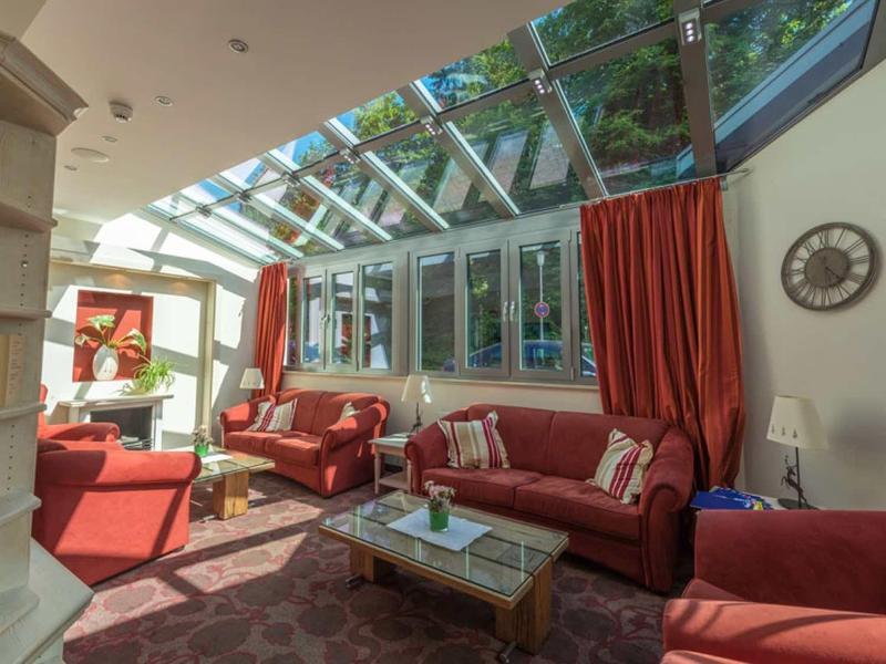 Helles Wohnzimmer mit roten Möbeln, großen Fenstern und Glasdach, Teppich und Bücherregal.