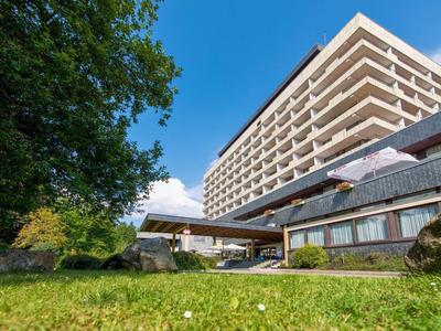 Moderní hotelová budova s balkony, obklopena zelení a modrou oblohou.