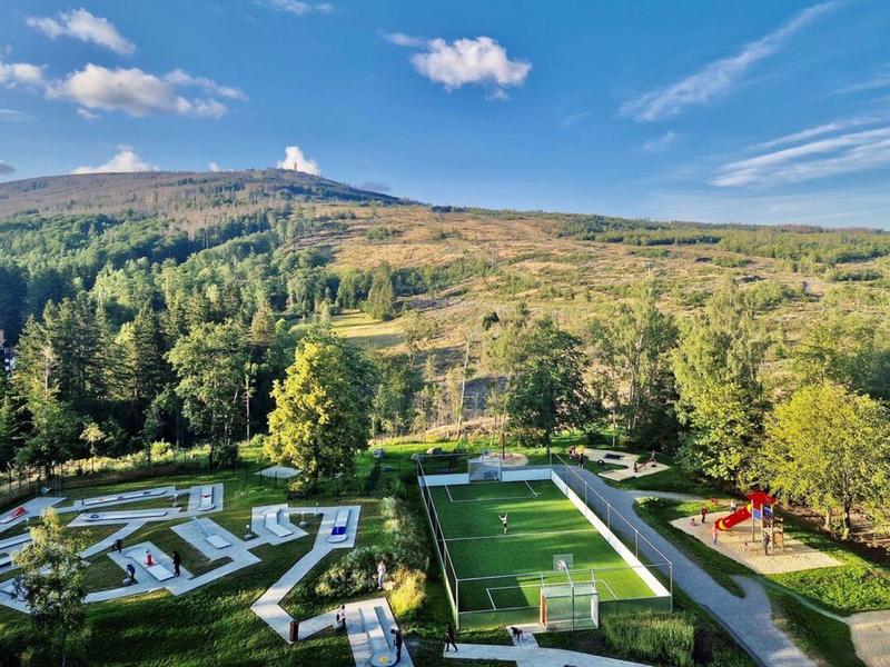Parkgebied met midgetgolf, voetbalveld en speeltuin voor beboste heuvel onder blauwe lucht.