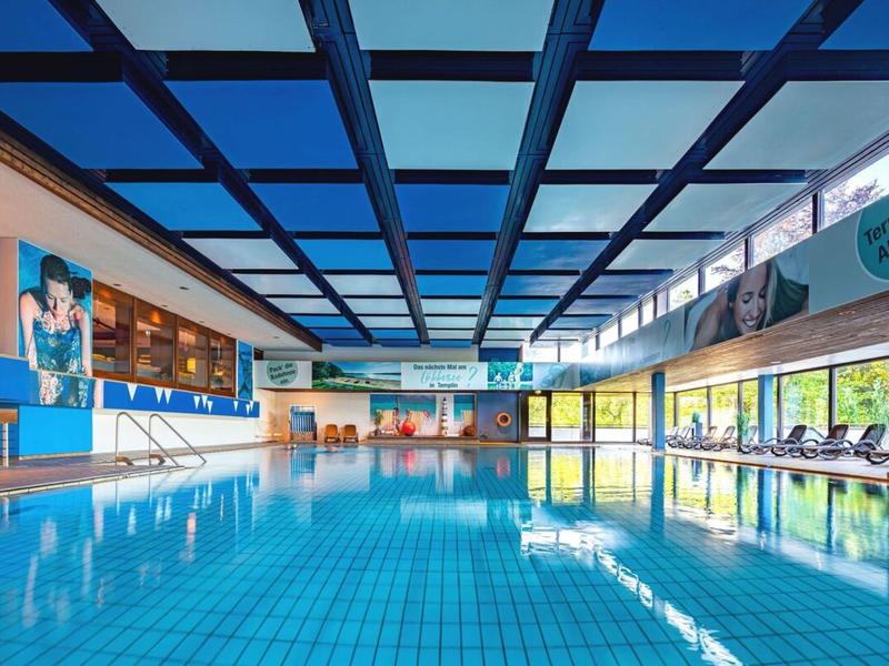 Licht overdekt zwembad met blauwe plafond en grote ramen, ligstoelen naast het bad