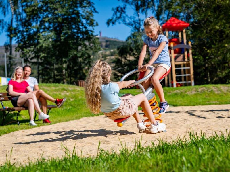 Kinderen spelen op een schommel buiten terwijl volwassenen toekijken in een zonnig park.