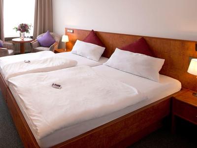 Ein schlicht eingerichtetes Schlafzimmer mit zwei Einzelbetten, Nachttischlampen und Sitzbereich am Fenster.