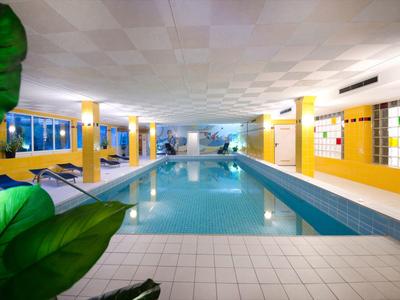 Hallenbad mit langem rechteckigem Pool, gelben Wänden und weißen Fliesen, grüne Pflanzen im Vordergrund.