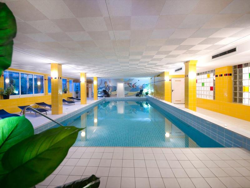 Hallenbad mit langem rechteckigem Pool, gelben Wänden und weißen Fliesen, grüne Pflanzen im Vordergrund.