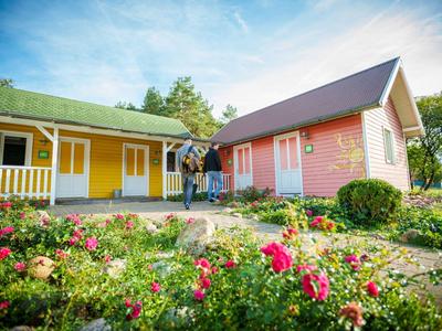 Zwei Personen stehen vor farbigen Holzhütten mit einem Blumengarten im Vordergrund.